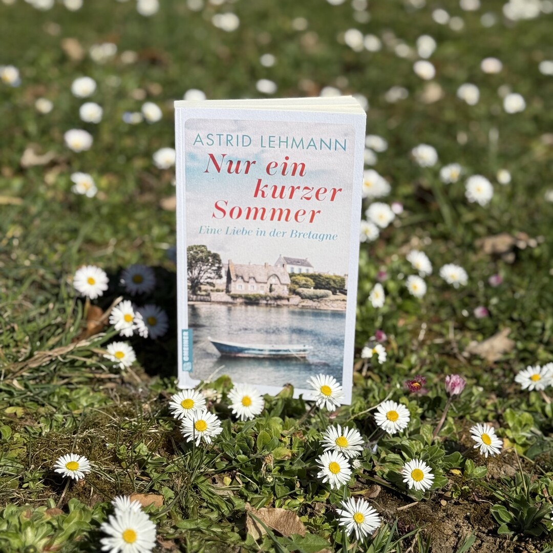 Auf einer gr&uuml;nen Wiese mit G&auml;nsebl&uuml;mchen steht  das Buch "Nur ein kurzer Sommer" von Astrid Lehmann. Das Cover des Buches zeigt ein Ruderboot auf einem See, im Hintergrund ist ein altes Stein-Haus zu sehen.