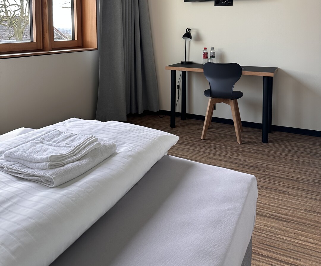 Zimmer am Mediacampus Frankfurt. Links befindet sich ein Fenster mit einem grauen Vorhang, der beiseite geschoben wurde. Das Bett hat wei&szlig;e Bettw&auml;sche und auf dem Bett liegen zwei wei&szlig;e, gefaltete Handt&uuml;cher. An der Wand gegen&uuml;ber dem Bett steht ein Schreibtisch mit einer Schreibtischlampe und zwei Flaschen Wasser. Davor steht ein Stuhl.
